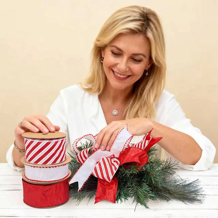 A woman crafts a Christmas decor with red-white striped, pom-pom-trimmed, and solid red ribbons on green pine.

