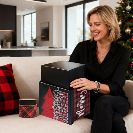 Woman with red-black plaid gift box (with "Be Merry" and Christmas tree) and red-black plaid ribbon spool, enjoying unboxing by a Christmas tree.