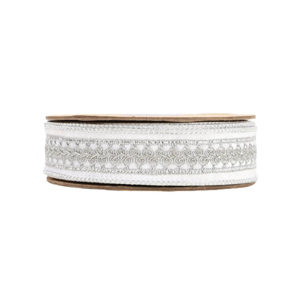 A roll of ribbon displays an elegant pattern of silver embroidery and metallic accents on a white base.