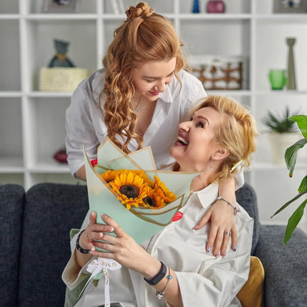 Young woman gifting a sunflower bouquet wrapped in sage matte floral sheets with gold edge to an older woman