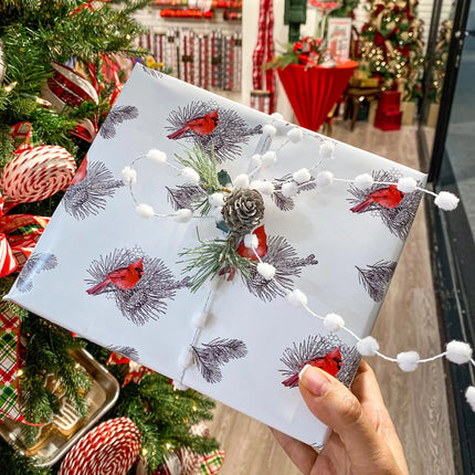 Christmas gift wrapped in holiday cardinal pinecone wrapping paper, decorated with pom pom garland and pinecone