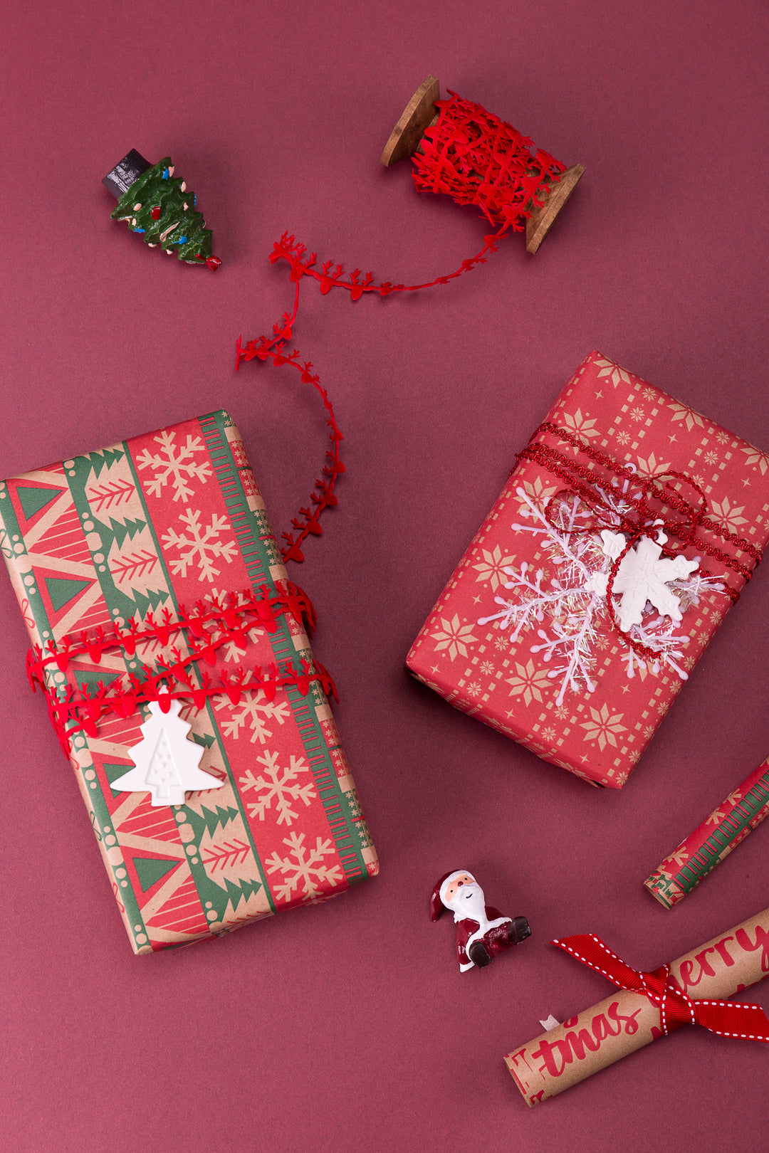 Red kraft gift boxes on a maroon table, next to a red gift ribbon spool